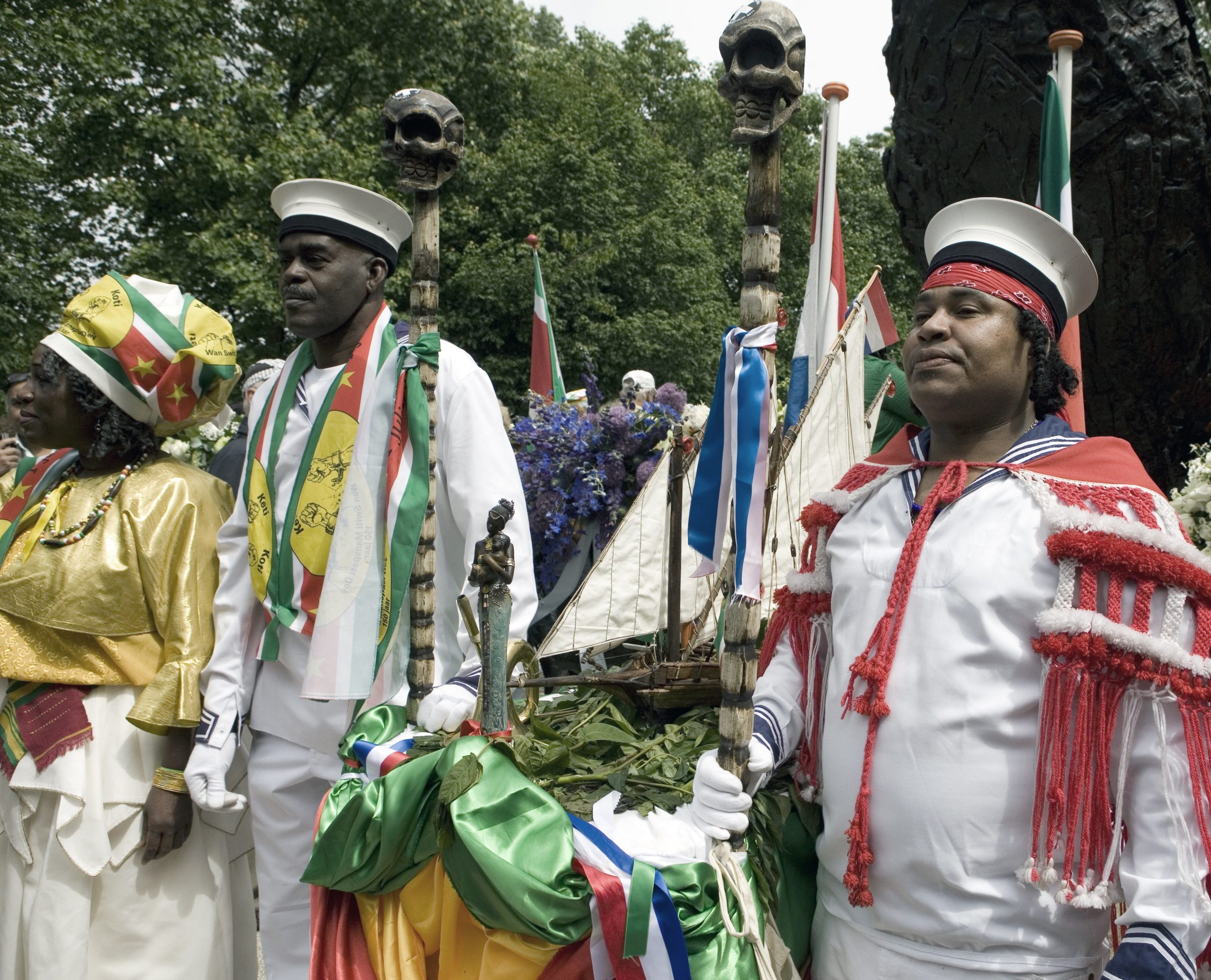 Groepsportret van mannen in traditionele kledij die een schip model dragen bij het nationaal monument slavernijverleden in het Oosterpark op 1 juli 2013. Bron: Stadsarchief Amsterdam, CCO, vervaardiger Ton van Rijn.